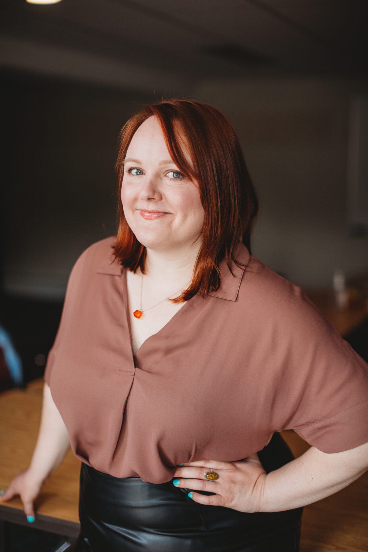 Amy Huziak — Indoor Headshot Amy stands smiling with one hand on her hip in a boardroom.